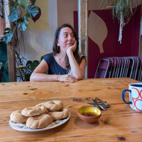 A woman with long brown hair and hoop earrings sits at a wooden table, resting her chin on her hand. On the table are bread rolls, a small bowl of dip, stacked spoons, and a mug with a red pattern. Plants decorate the background.