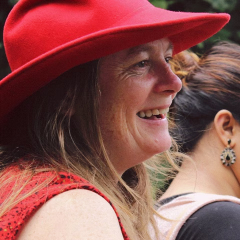 A woman with long hair smiles, wearing a wide-brimmed red hat and a red top. Another person with earrings and dark hair in a bun is partially visible in the background.
