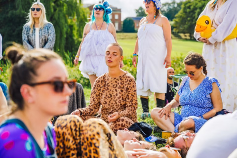 A group of people, some in colourful or animal-themed outfits, gather outdoors. One person in a leopard-print suit sits cross-legged with eyes closed, as others stand or sit nearby, some wearing sunglasses. The mood is relaxed and peaceful.
