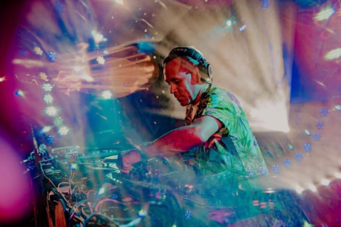A DJ wearing headphones and a colourful shirt mixes music at a console, surrounded by vibrant, multi-coloured lights and dynamic visual effects creating an energetic party atmosphere.
