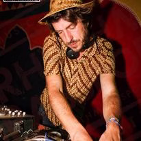 A DJ wearing a patterned shirt and straw hat is mixing music on turntables, focused on his equipment, with a red and black background behind him.