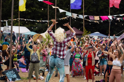 A performer with a curly blonde wig and checked shirt leads a lively outdoor crowd, who are raising their hands and dancing. Colourful flags and bunting decorate the festival scene under a sunny sky.