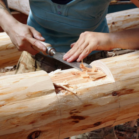 A person uses a chisel and mallet to carve and shape a large wooden log, with wood shavings curling away from the tool. The person is wearing a blue shirt and camouflage-patterned trousers.