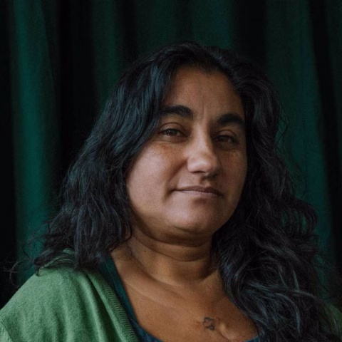 A woman with long, dark, wavy hair and medium skin tone, wearing a green jumper, stands in front of dark green curtains, looking slightly to the side with a gentle expression.