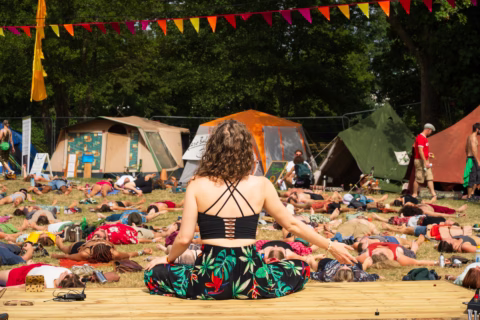 A woman sits cross-legged on a wooden platform, leading a group yoga or meditation session outdoors. Participants lie on the grass surrounded by tents, under colourful bunting and trees.