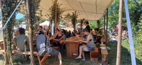 A group of people sit at wooden tables under a canopy decorated with branches, attending a workshop or talk at an outdoor festival on a sunny day. Trees and other marquees are visible in the background.