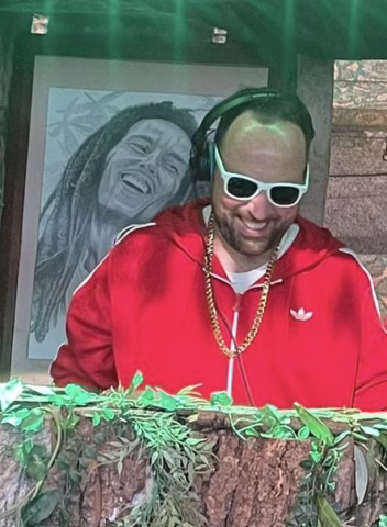 A man in a red tracksuit, white sunglasses, and a gold chain smiles while wearing headphones. Behind him is a drawing of Bob Marley.