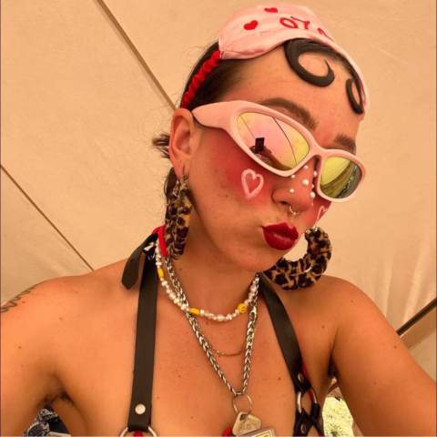 A person wearing pink sunglasses, a pink hairband, bold red lipstick, heart face paint, and statement earrings poses for a selfie under a marquee. They have unique facial make-up and accessories, including layered necklaces.
