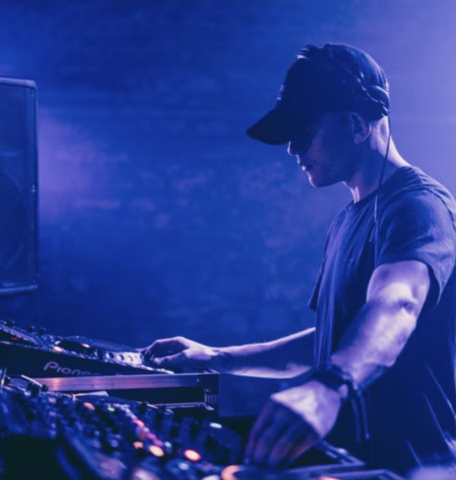 A DJ wearing a black cap and headphones mixes music on a mixing desk in a dimly lit, blue and purple-toned setting.
