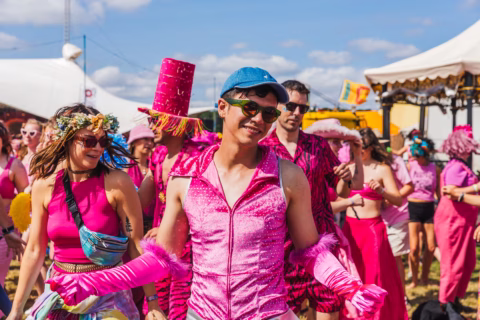 A group of people dressed in vibrant pink costumes, some with hats and gloves, enjoying an outdoor festival on a sunny day with marquees and decorations in the background.