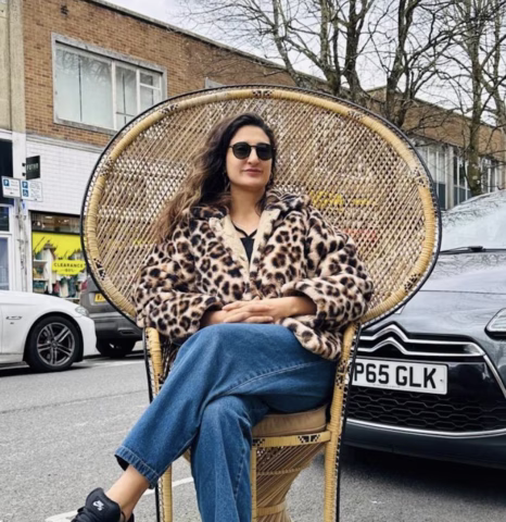 A woman wearing sunglasses, a leopard print coat, and blue jeans sits cross-legged on a wicker chair placed on a street, with parked cars and shopfronts in the background.