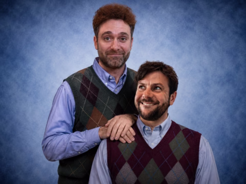 Two men wearing argyle jumper vests and blue shirts pose together in front of a blue textured backdrop, with one man standing and resting his hands on the seated man's shoulders. Both are smiling.