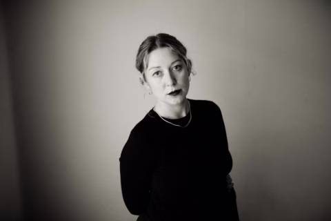 A woman with fair hair tied back, wearing a dark long-sleeved top and a simple necklace, stands in front of a plain wall. She looks directly at the camera with a neutral expression in a black-and-white photo.