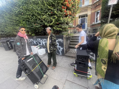 Four people stand on a city pavement with portable speakers and equipment. The background features a graffiti-covered wall, greenery, and a brick building. One person wears a green headscarf, and others wear casual clothing and sunglasses.