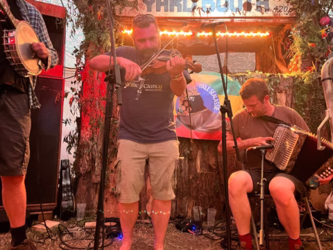 Three musicians perform on a rustic stage. The central man plays a violin, another to the right sits playing an accordion, and the third on the left holds a banjo. Warm lights and greenery decorate the background.