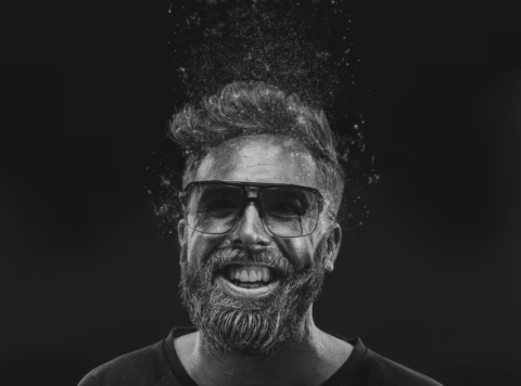Black and white photo of a bearded man wearing sunglasses, smiling widely. His hair and beard appear to be dissolving or transforming into particles, blending into a dark background.