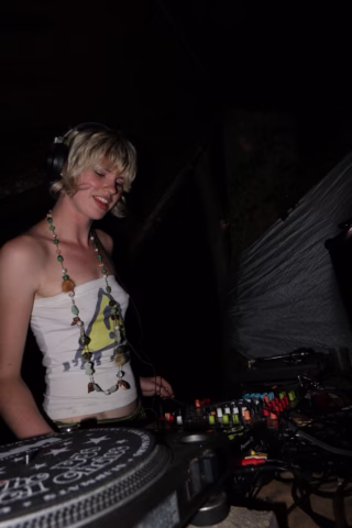 A woman with short blonde hair wearing headphones and a vest stands behind DJ equipment, smiling as she mixes music in a dimly lit setting.