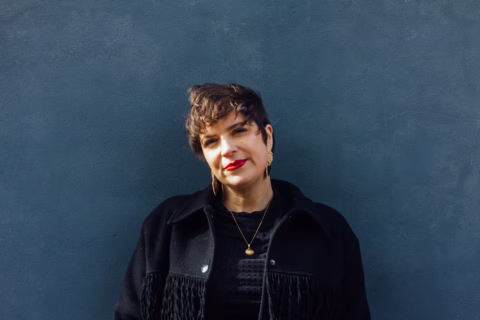 A person with short brown hair and red lipstick, wearing a black fringed jacket and gold necklace, stands in front of a plain dark blue wall, smiling slightly.