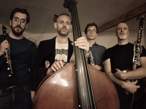 Four men stand indoors holding musical instruments, including clarinets and a double bass, and look seriously at the camera. The background features a beige wall and a wooden beam.