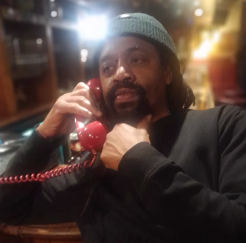 A man wearing a dark jumper and green knitted hat sits at a bar, holding a red rotary telephone to his ear and looking thoughtful. The background is softly blurred with warm lighting.