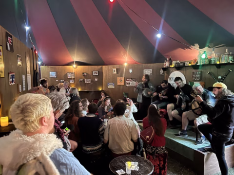 A lively group of people gather in a dimly lit circus-style tent bar, some playing instruments and singing on a small stage whilst others sit, talk, and enjoy drinks around tables. The décor includes photos, lights, and bottles on shelves.
