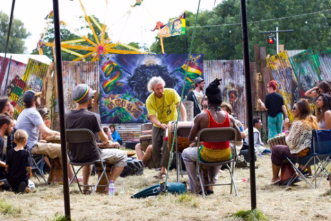 A group of people sit in a circle outdoors, playing drums at a festival. Colourful artwork, funfair rides, and signs are in the background, with trees and fairy lights overhead.