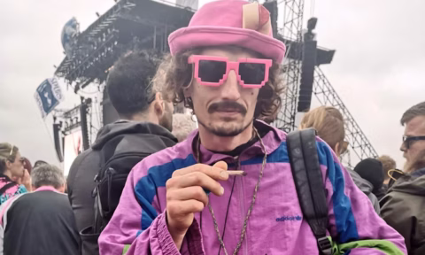 A man in a purple retro jacket, pink pixelated sunglasses, and a pink hat holds a rolled cigarette at an outdoor music festival, with a stage and crowd visible in the background.