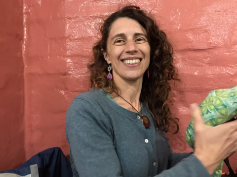 A woman with curly brown hair, wearing a blue cardigan and colourful earrings, smiles whilst sitting in front of a pink textured wall. She is holding a green patterned item in her hand.
