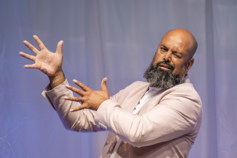 A bearded man in a light-coloured suit gestures expressively with his hands and squints one eye, standing against a plain, softly lit background.