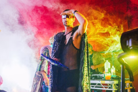 A person wearing futuristic sunglasses, a black mesh outfit, and a dramatic fur cape stands onstage in colourful, smoky lighting, holding a shiny fan and posing confidently. Other people and equipment are in the background.