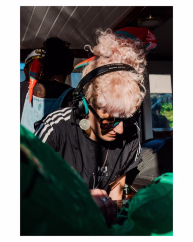 A person with curly, light-coloured hair, headphones, and sunglasses is DJing at a sunlit event, focused on the equipment. They wear a black shirt with white stripes and a necklace, with party decorations in the background.