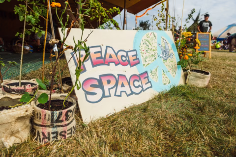 A painted sign on grass reads “PEACE SPACE” in colourful letters, with a peace symbol, dove, and leaves. Small potted plants in sacks surround the sign, and people are seated under a nearby marquee.