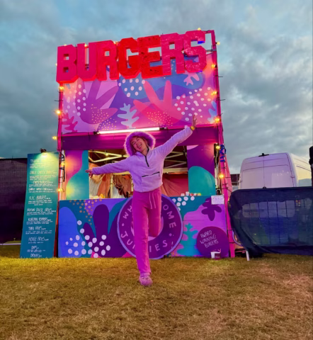A person in a pink outfit and hoodie jumps joyfully in front of a colourful burger stall with a large BURGERS sign, surrounded by festival decorations and fairy lights at dusk.