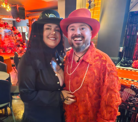 A smiling couple dressed in vibrant costumes pose indoors; the woman wears a black veil and hat, whilst the man wears a bright red furry suit, hat, and beaded necklaces. The festive background features red and black decorations.