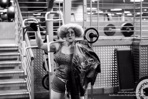 A person with curly hair, round glasses, and dramatic make-up stands in a gym holding a jacket that says NA NA RACIST. They lean on gym equipment, looking at the camera. The scene is in black and white.