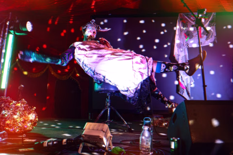 A performer in a whimsical costume appears to float horizontally on stage, surrounded by colourful lights and disco-style dots, with stage equipment and props visible in the foreground.