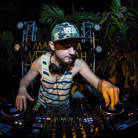 A DJ wearing a striped vest, headphones around his neck, and a camouflage cap mixes music on a DJ controller at an outdoor event, surrounded by palm trees and stage lights.