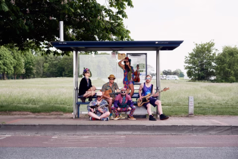 A group of six people in colourful, eclectic clothing pose at a bus stop on a rural roadside; some are seated, some standing, with musical instruments and a grassy field in the background.