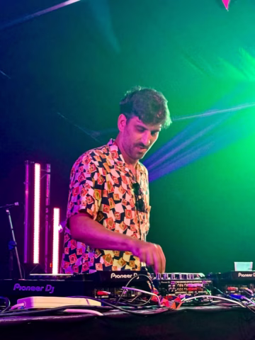 A DJ wearing a colourful patterned shirt stands behind a Pioneer DJ controller, focused on mixing music under vibrant green and purple stage lights.