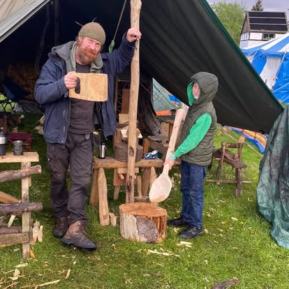 A man and a child stand by a canvas tent outdoors. The man smiles and holds a wooden mug, while the child in a green hoodie and waistcoat holds a large wooden spoon, both near rustic wooden stools and chopping blocks.