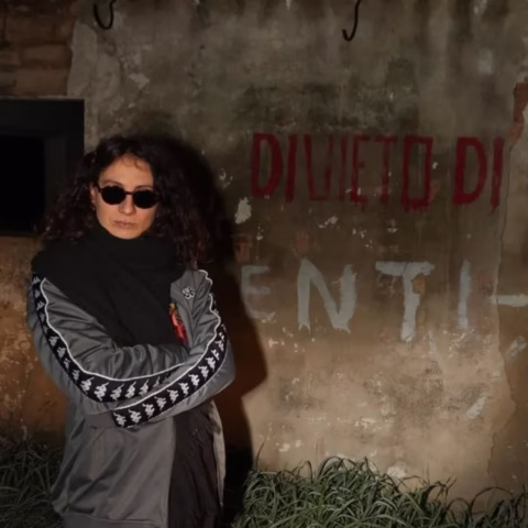 A person with curly hair, wearing dark sunglasses and a grey jacket with patterned sleeves, stands in front of a weathered wall with faded red and white writing that partially reads DIVIETO DI. There is grass at the bottom of the wall.