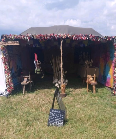 A colourful festival marquee decorated with fabric strips and banners stands on grass. Inside are wooden chairs, tables, and craft displays. A chalkboard sign with handwritten text is positioned outside the entrance.