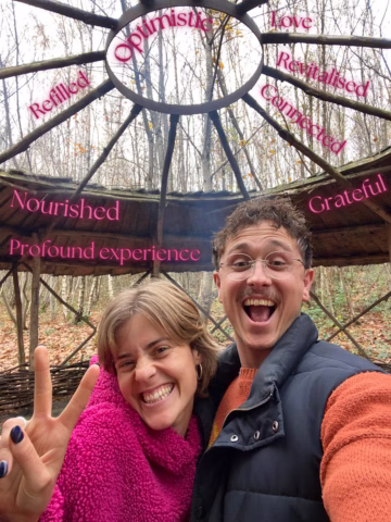 Two smiling people take a cheerful selfie under a wooden structure in the woods. Words like Optimistic, Grateful, and Nourished are written in pink above them. One person flashes a peace sign.