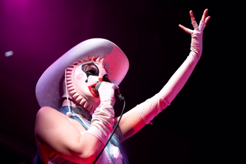 A performer wearing a dramatic white hat, ruffled mask with painted red lips and cheeks, and long white gloves sings into a microphone under pink stage lighting with one arm raised.