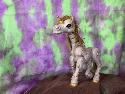 A wooden unicorn figurine with gold hooves and a curly gold mane stands on a brown surface against a purple and green tie-dye background.