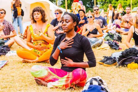 A diverse group of people sits cross-legged on grass, eyes closed, with one hand on their chest and one on their abdomen, appearing to meditate or practise mindful breathing at an outdoor event or festival.