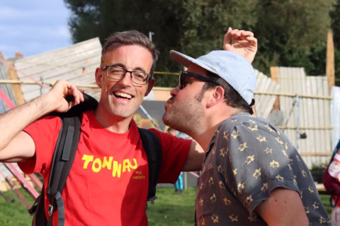 Two men smiling outdoors; one in a red shirt and glasses with a rucksack, the other in a patterned shirt and cap playfully leaning in as if to kiss the first man's cheek. Festival-like setting in the background.