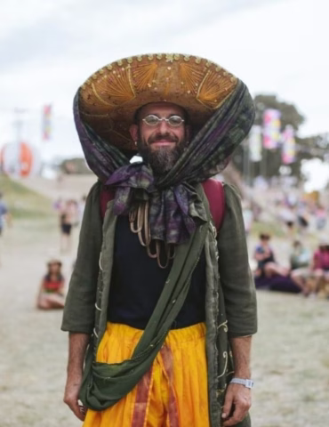 A smiling man outdoors wears a large, ornate hat, round glasses, a bulky green scarf tied in a bow, a dark shirt, and bright yellow trousers. Blurry festivalgoers and colourful flags are visible in the background.