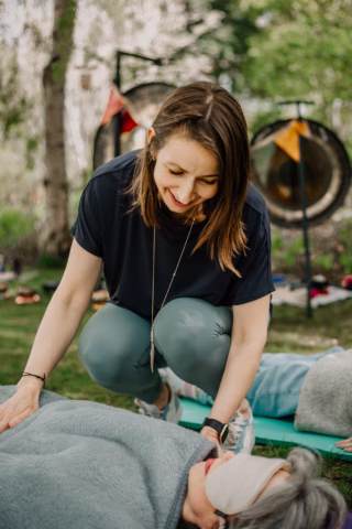 A woman kneels on the grass outdoors, smiling as she gently touches a person lying down covered with a blanket and wearing an eye mask. Gongs and colourful flags are visible in the background.