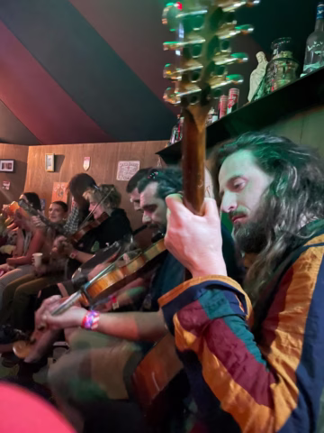 A group of people sit closely together indoors, playing stringed instruments. The focus is on one man in a colourful jacket, holding a guitar. The background is filled with more musicians, wooden walls, and various decorations.